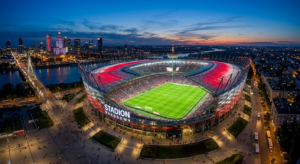 A professional wide-angle aerial shot of a modern, floodlit Polish football stadium under a twilight